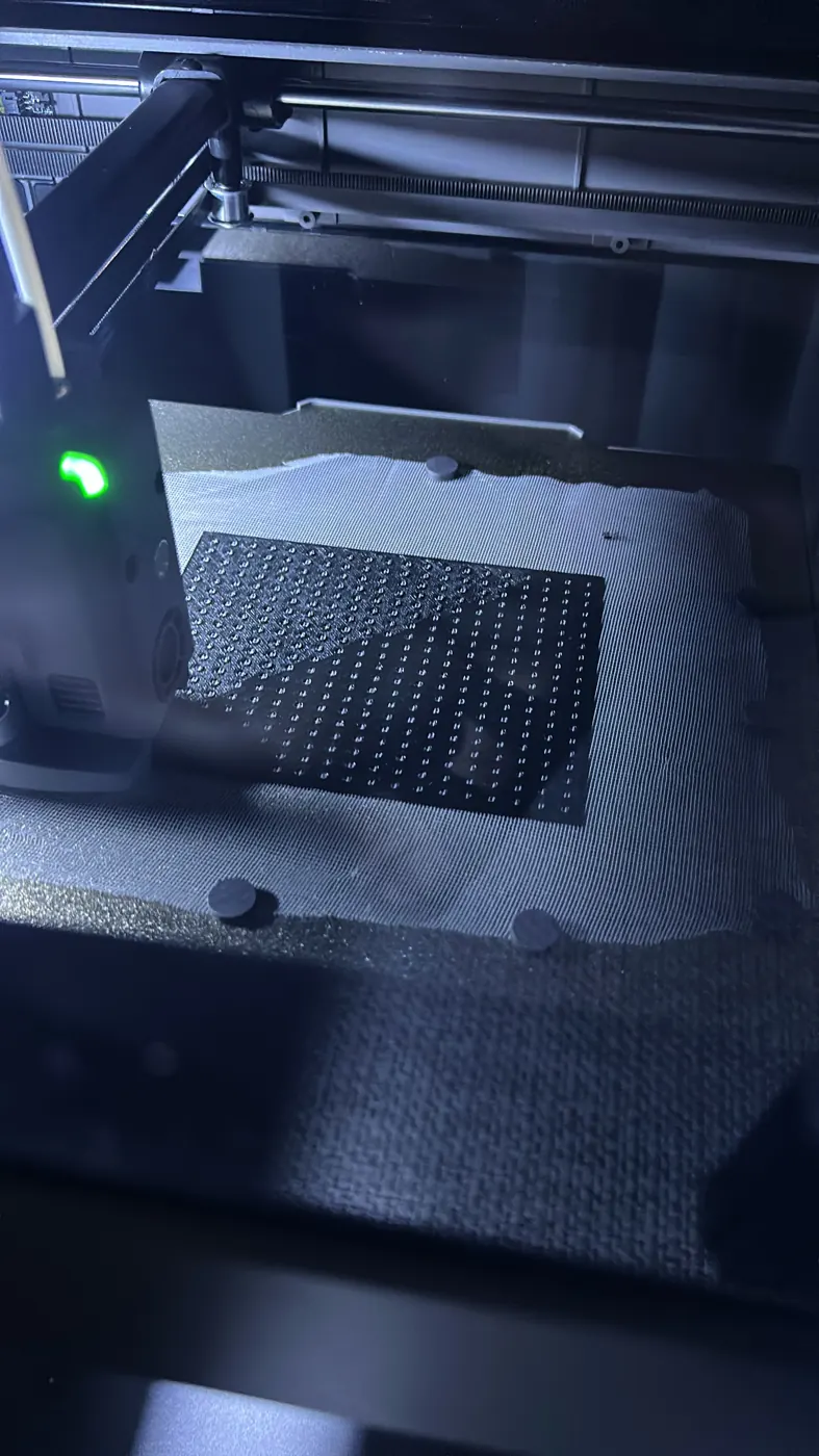 3D yazıcı tablasına küçük ağırlıklarla sabitlenmiş ince tül üzerinde basılan, düzenli deliklerle kaplı siyah dikdörtgen hoparlör kapağı