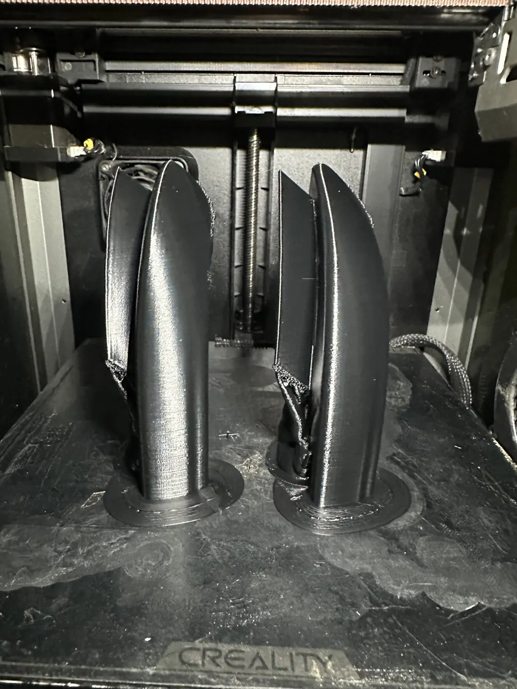 Kapalı kasalı 3D yazıcının tablasında dik olarak basılmış iki kavisli trim parçası, alt kısımlarında ağaç destek ve brim